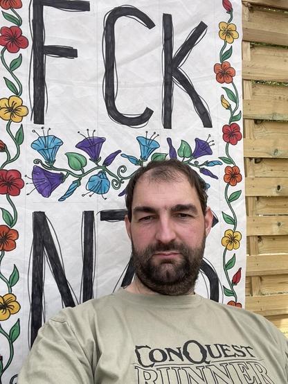 Foto von mir  vor einem blumigen FCK NZS Banner, ich trage ein „Conquest Runner“ T-Shirt