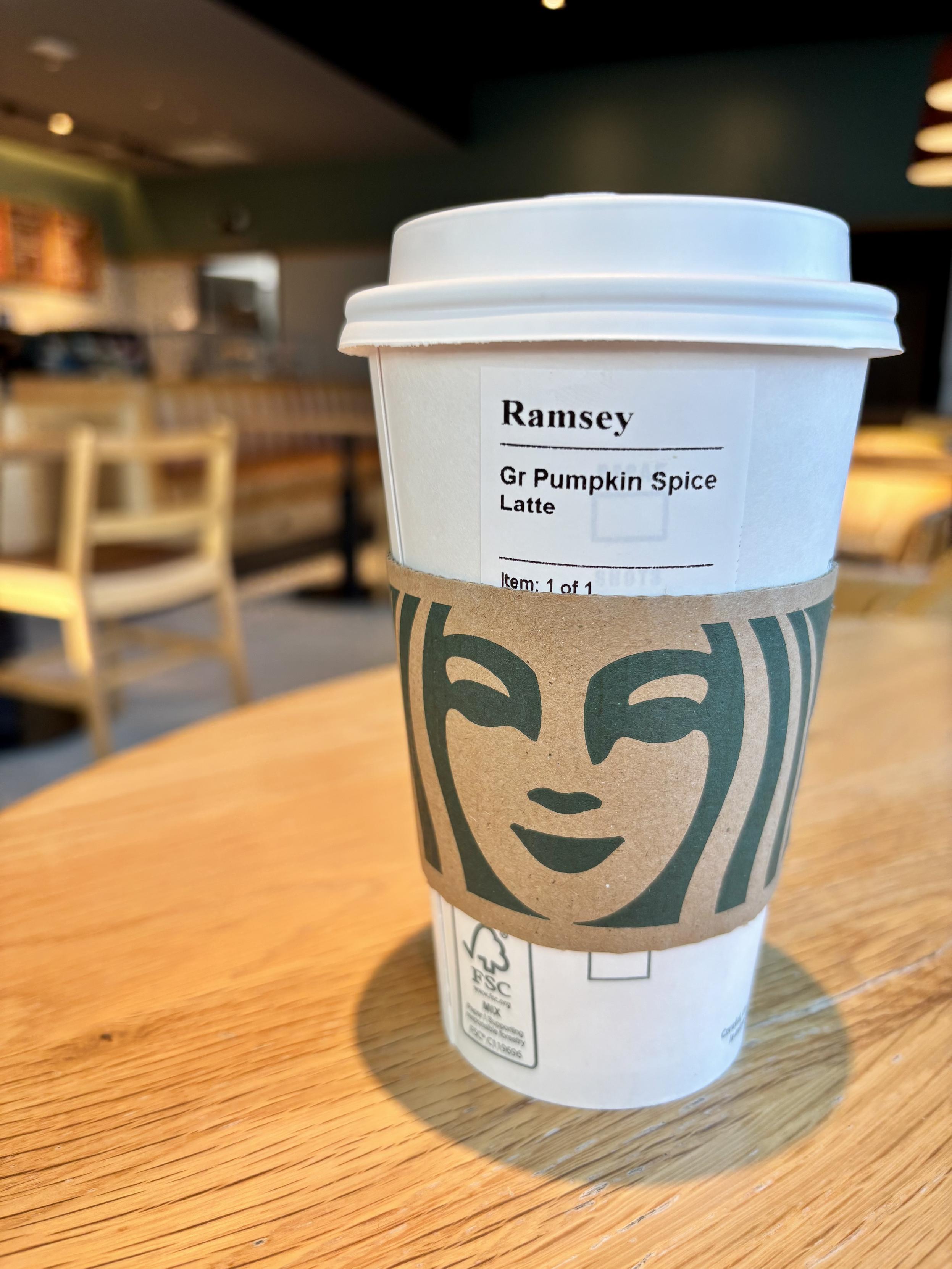 A Starbucks cup with cardboard sleeve is on a table in the foreground of the image. The blurred background is the interior of a Starbucks cafe. The label on the cup reads: “Ramsey. Gr Pumpkin Spice Latte.”