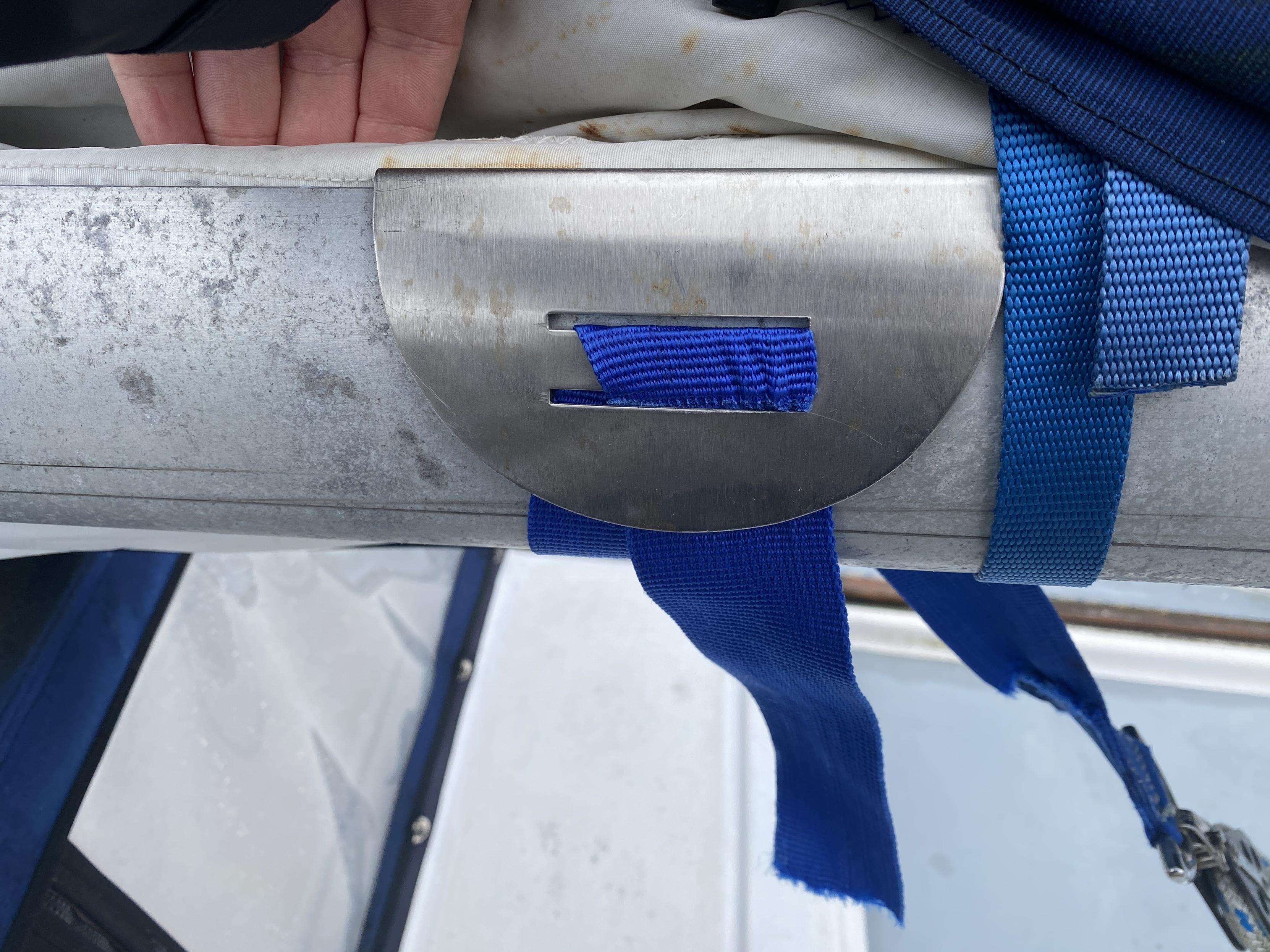 A metal clip fitted between the boom and the sail, with a blue mesh strap fed through two slots. The frayed end of the strap can be seen near the bottom.