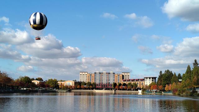 A hotel across the water with a hot air balloon floating to the left of it. The sky is blue with a few fluffy clouds.