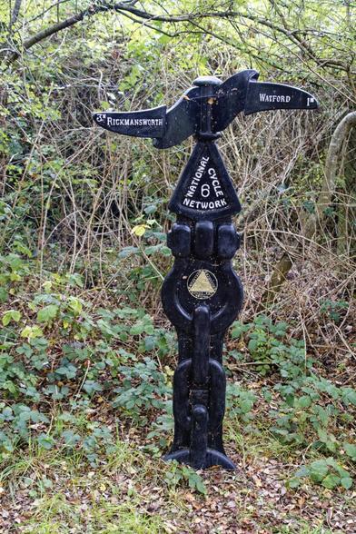 A fancy black pillar made out of fake chains. It has two pointers. One to Rickmansworth, and one to Watford.