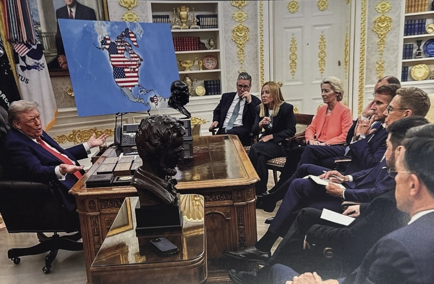 Photo of Trump in the oval office with a map on display that originally showed Ukraine but was doctored by Trump to show North America with Canada, Greenland and Venezuela covered in an American flag. Other officials in the room are sitting calmly and listening to him.
