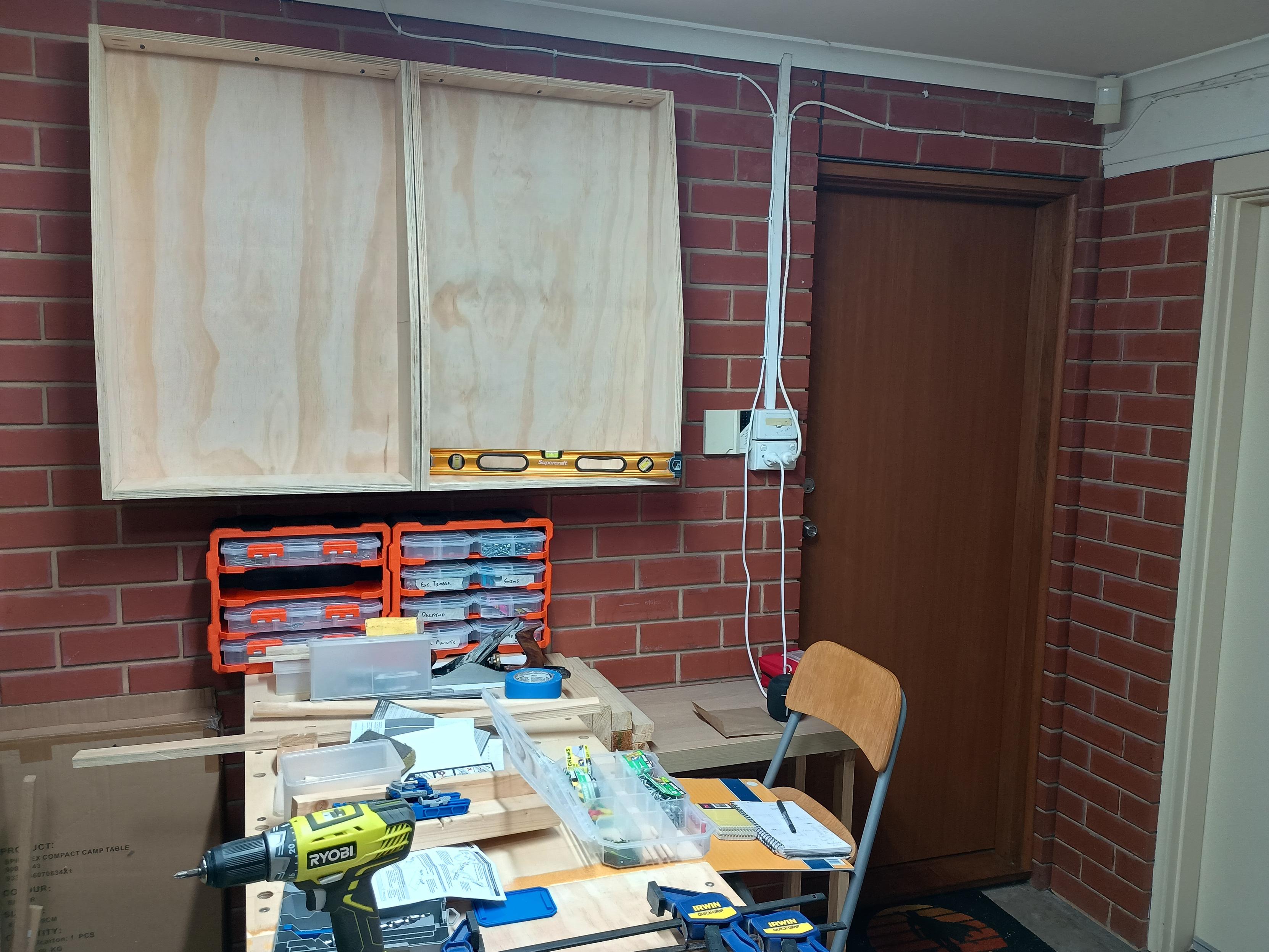 A woodwork hop. Two empty hollow boxes have been hung on the wall above a long workbench which is perpendicular to the wall, allowing access to both long sides of the workbench.

The work bench is covered in tools and offcuts of wood.