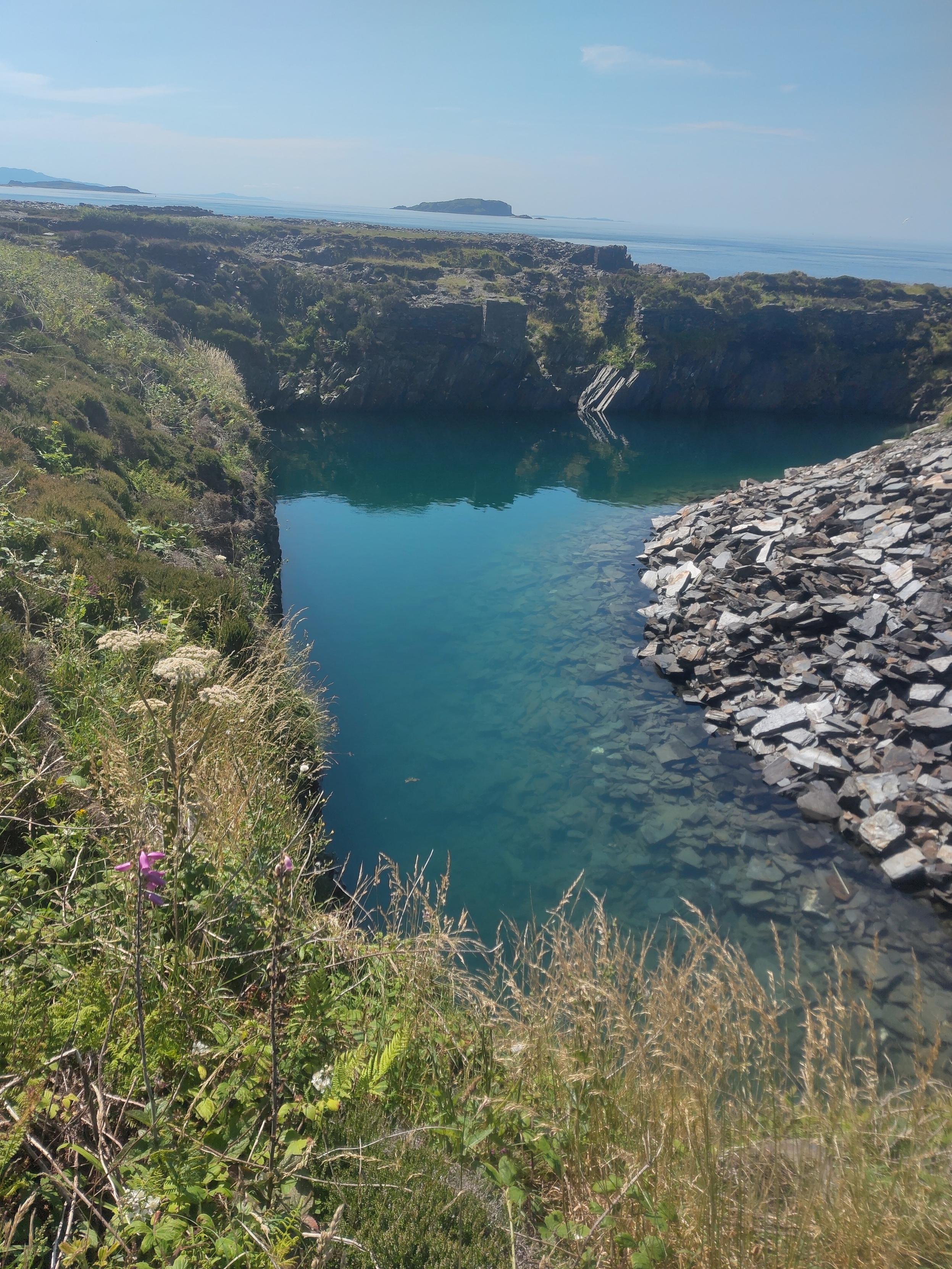 A former.slate quarry that is now full of water. The water is rather clear so the slate debris can be seen in the depth. In the background the Atlantic Ocean is visible above the rim.of the former.quarry. An island is visible some.10km afar