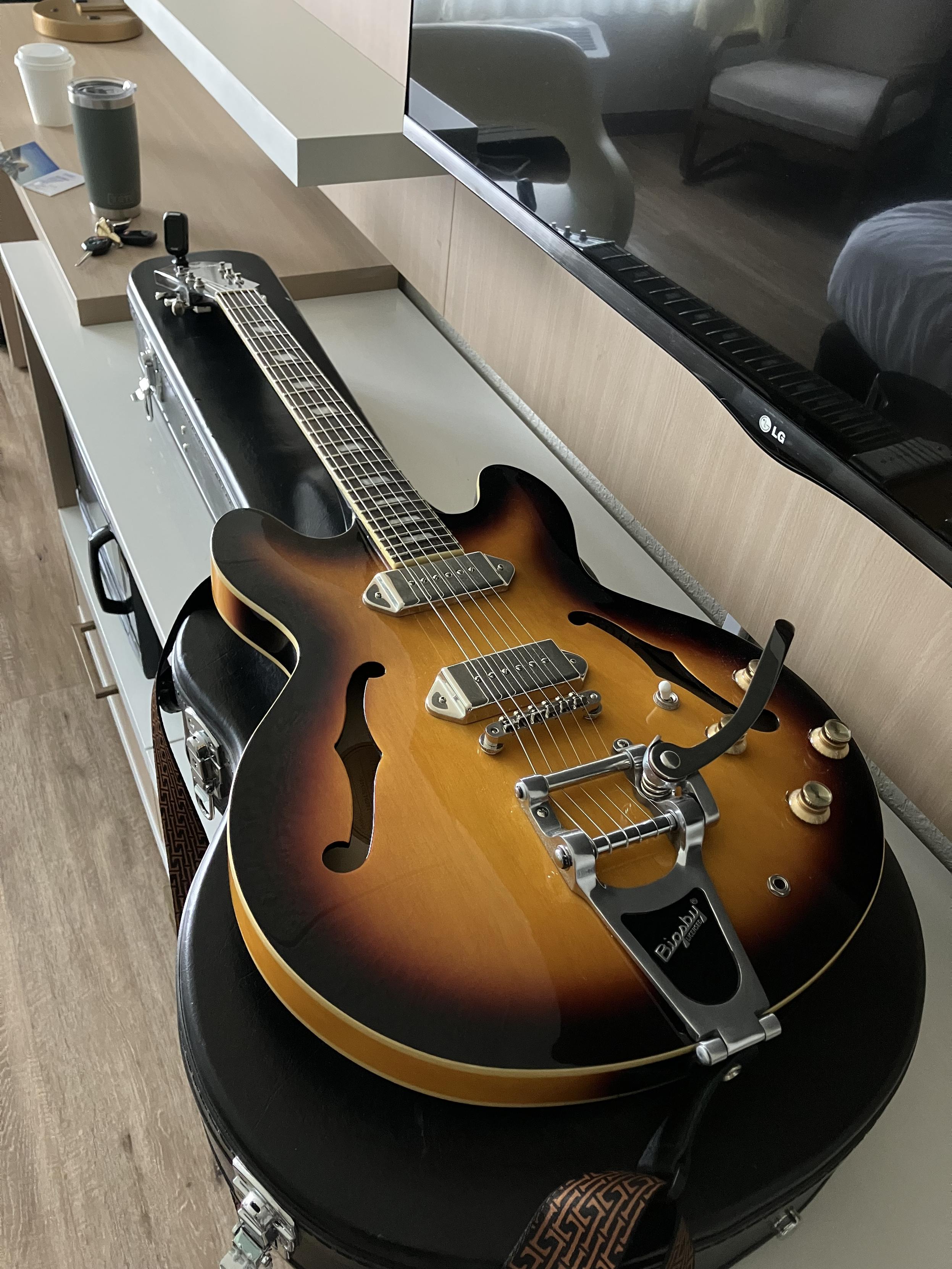 A sunburst Epiphone Casino with a Bigsby,lying on top of its case below a hotel TV