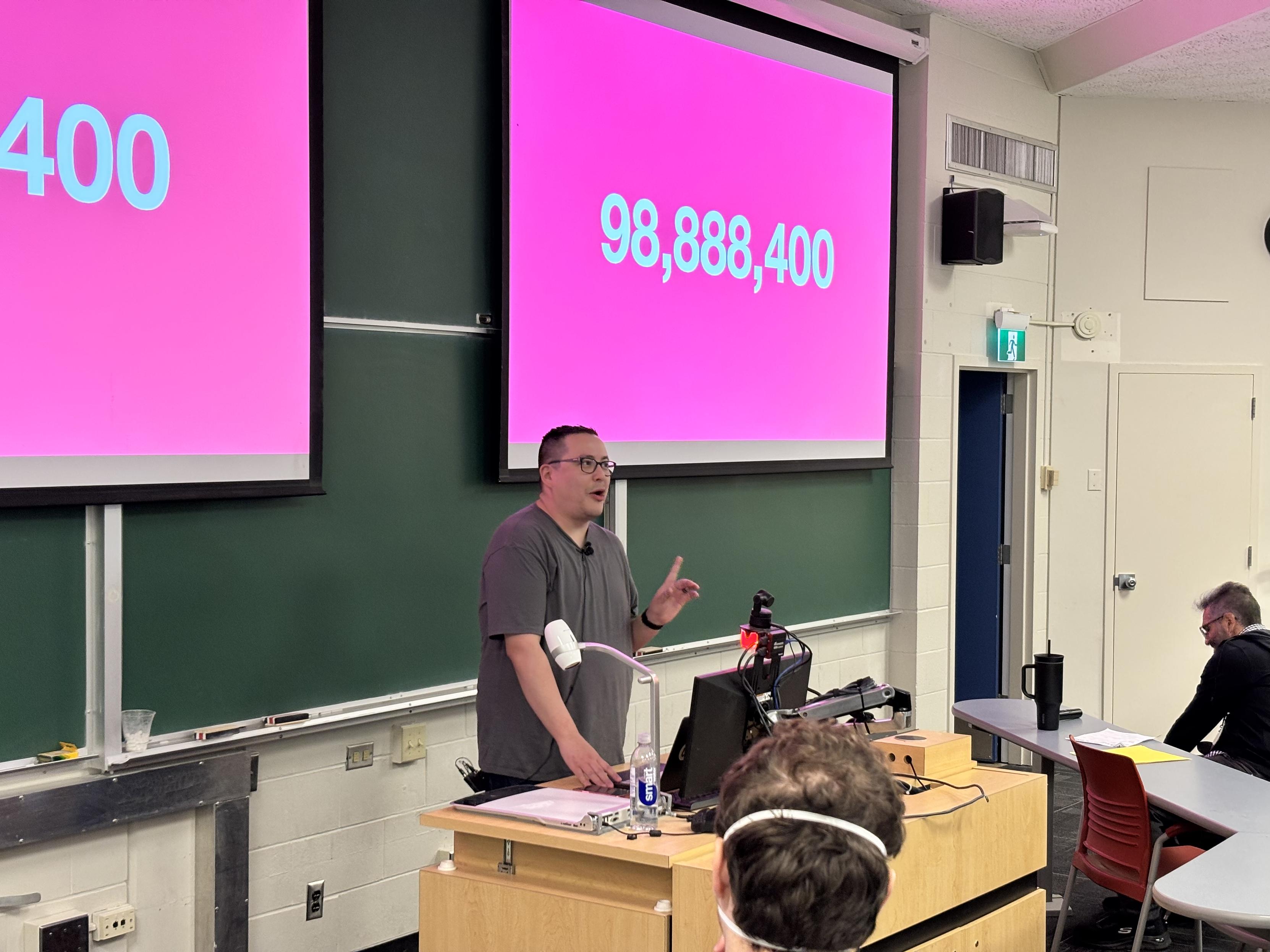 Daniel Supernault stands in front of a chalk board and a projection screen displaying a bright pink background and the number 98,888,400.