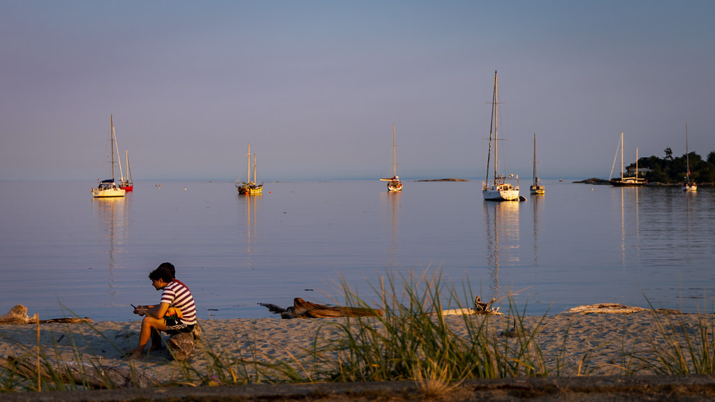Two young people sit on a log on the sandy beach at sunset. In the background, several sailboats are moored. The water is calm. Sunset/golden-hour light gives the scene a serene quietness.