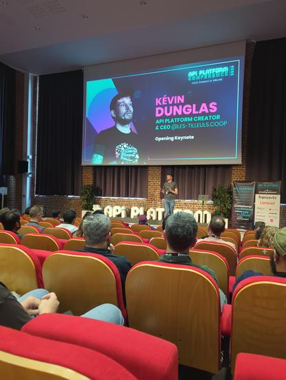 Ein Foto zeigt eine Konferenzveranstaltung in einem Auditorium. Eine große Projektionsfläche im Hintergrund zeigt das Porträt einer Person sowie den Text „API PLATFORM CONFERENCE 2023“, „LILLE (FRANCE) & ONLINE“, „KÉVIN DUNGLAS“, „API PLATFORM CREATOR & CEO [@]LES-TILLEULS.COOP“ und „Opening Keynote“. Vor der Leinwand steht eine Person auf einer Bühne. Im Vordergrund sind Reihen von roten Sitzen mit Holzrückenlehnen zu sehen, in denen ein Publikum von hinten sitzt. Auf der rechten Seite befinde…