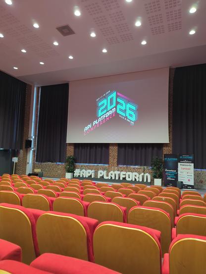 Ein leerer Konferenzsaal mit mehreren Reihen von Sitzplätzen, die auf eine große Projektionsfläche ausgerichtet sind. Die Sitze haben rote Polster und helle Holzrückenlehnen. Auf der Leinwand im Hintergrund ist eine Grafik mit dem Text „SEP 17-18 2026“, „2026“, „API PLATFORM CONFERENCE“ und „LILLE & ONLINE“ abgebildet. Unter der Leinwand befindet sich ein Banner mit der Aufschrift „#API PLATFORM“. An der rechten Seite stehen zwei vertikale Banner vor einer Ziegelwand, flankiert von dunklen Vorh…